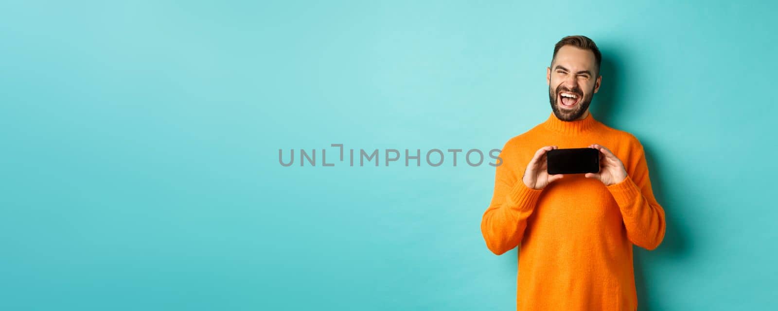 Photo of happy adult man laughing and showing funny thing on mobile phone screen, standing in orange sweater over turquoise background.