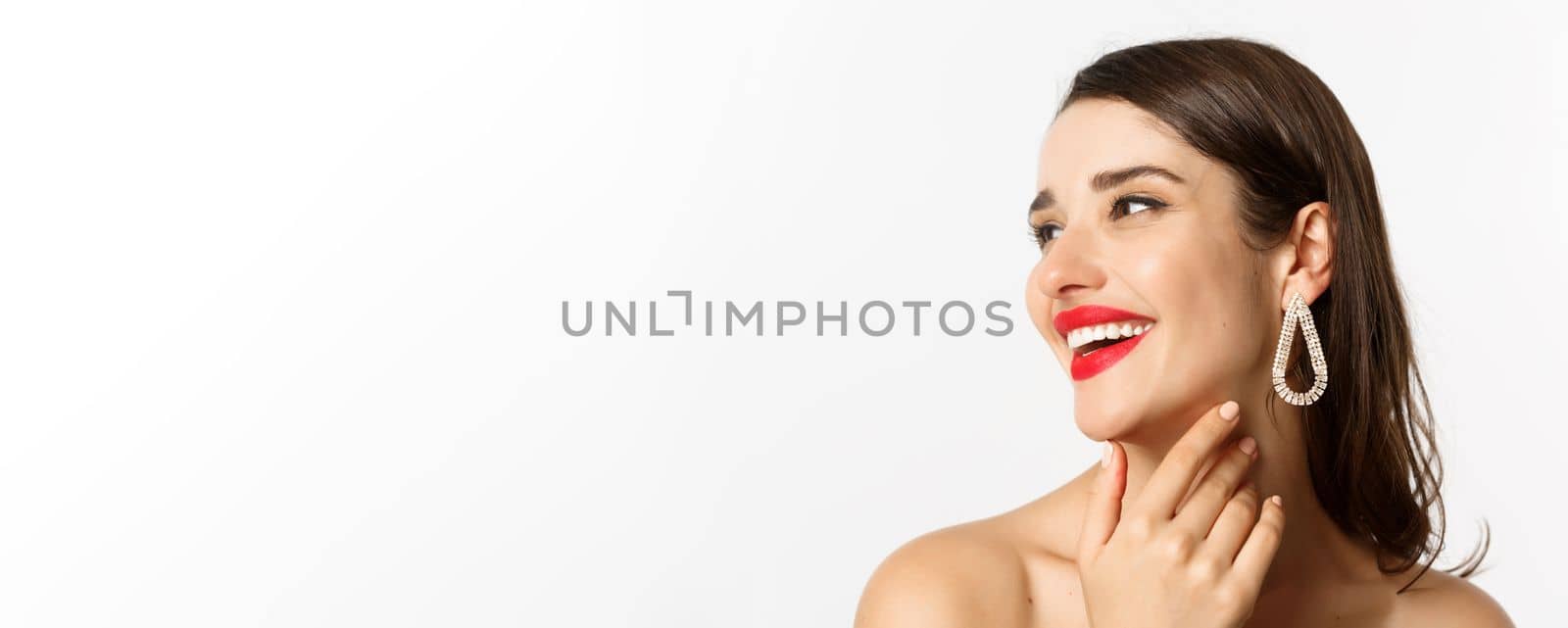 Fashion and beauty concept. Headshot of gorgeous brunette woman with red lipstick, earrings, laughing and looking left, standing over white background.