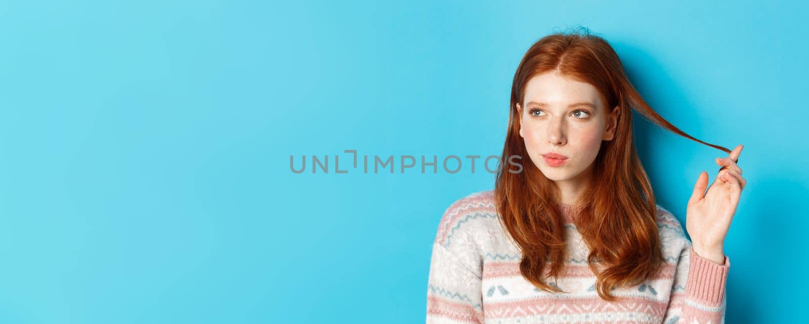 Close-up of thoughtful pretty redhead girl looking left, playing with hair strand and pondering, standing in winter sweater against blue background.