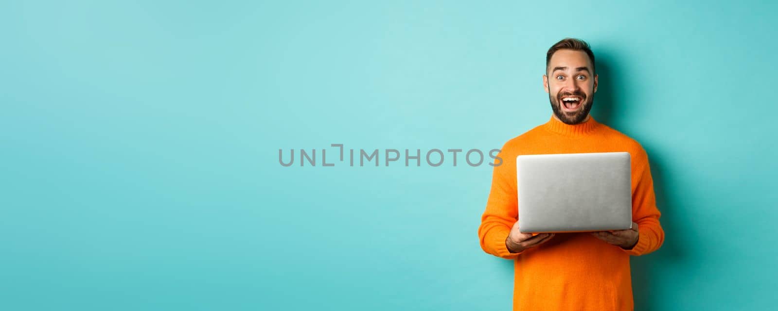 Happy man using laptop and looking excited at camera, standing with computer against light blue background.