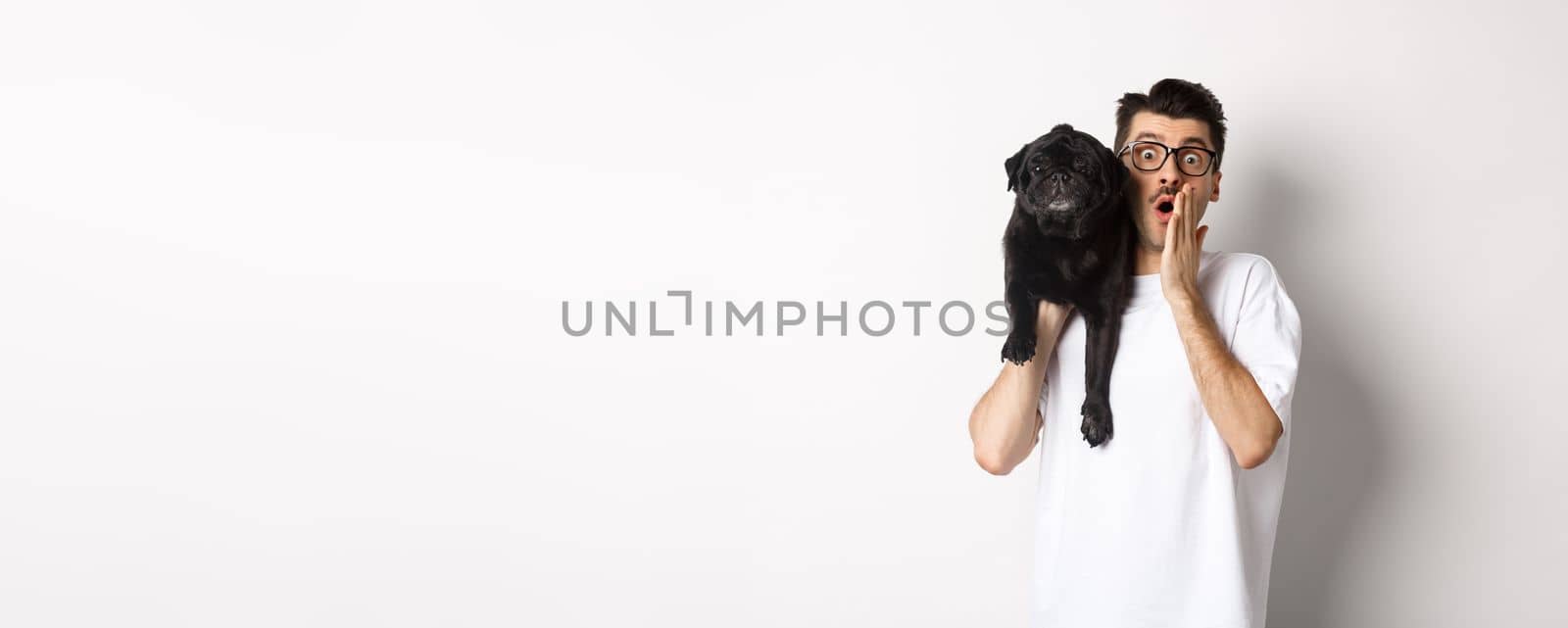 Image of shocked dog owner staring at camera and gasping impressed, cute black pug sitting on his shoulder and staring at you, white background.