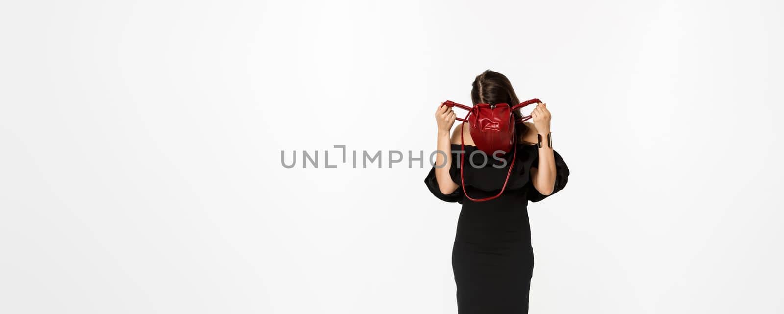 Beauty and fashion concept. Full length of young woman sticking head inside purse and searching something, wearing black dress and high heels, standing over white background by Benzoix