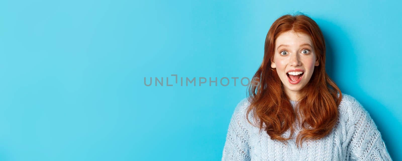 Close-up of surprised and excited redhead girl hear amazing news, staring at camera, looking at promo offer, standing over blue background by Benzoix