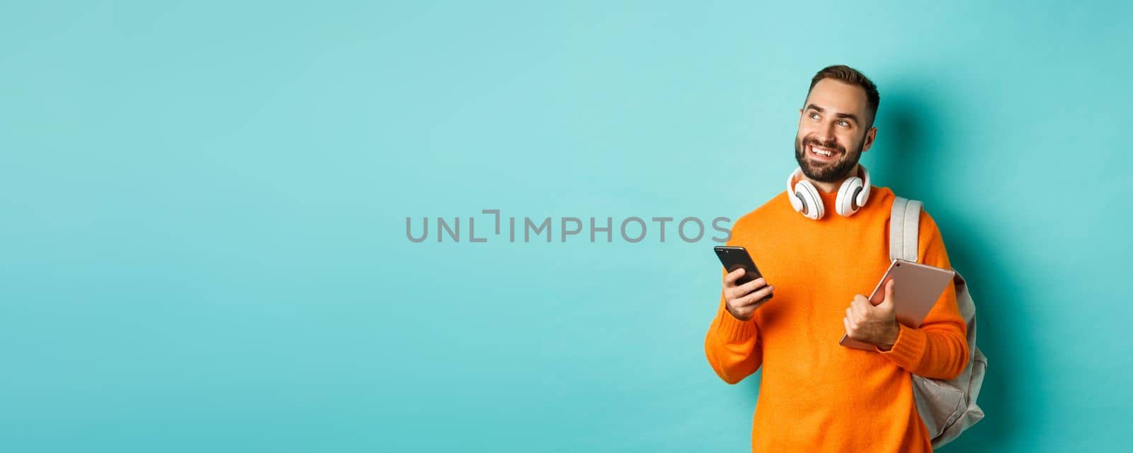 Handsome man student with headphones and backpack, holding digital tablet and smartphone, looking dreamy at upper left corner, standing against turquoise background.