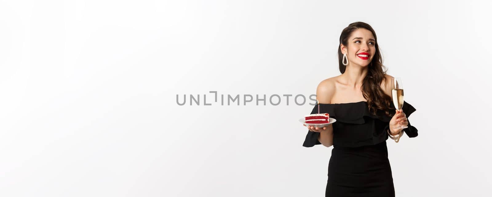 Celebration and party concept. Fashionable woman holding birthday cake with candle and drinking champagne, smiling and looking aside, standing over white background by Benzoix