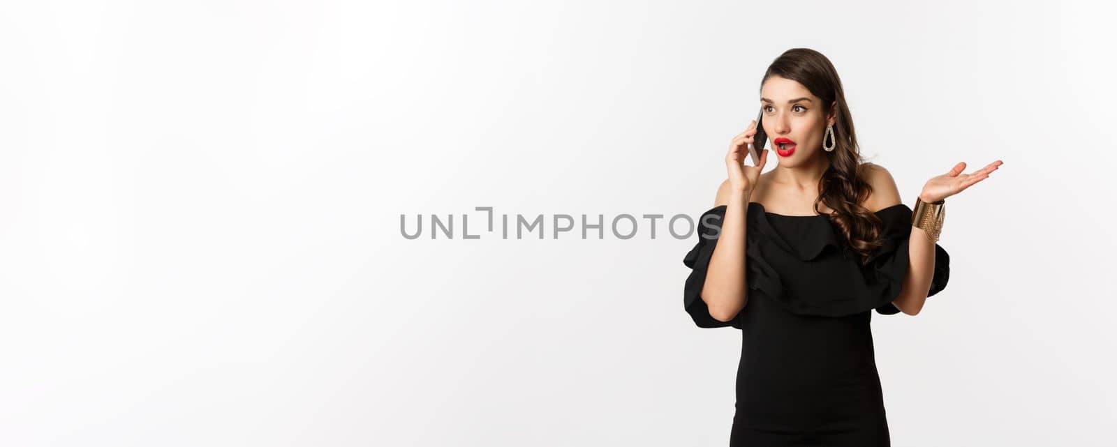 Attractive glamour woman in black dress talking on mobile phone, having conversation and looking surprised, standing over white background.