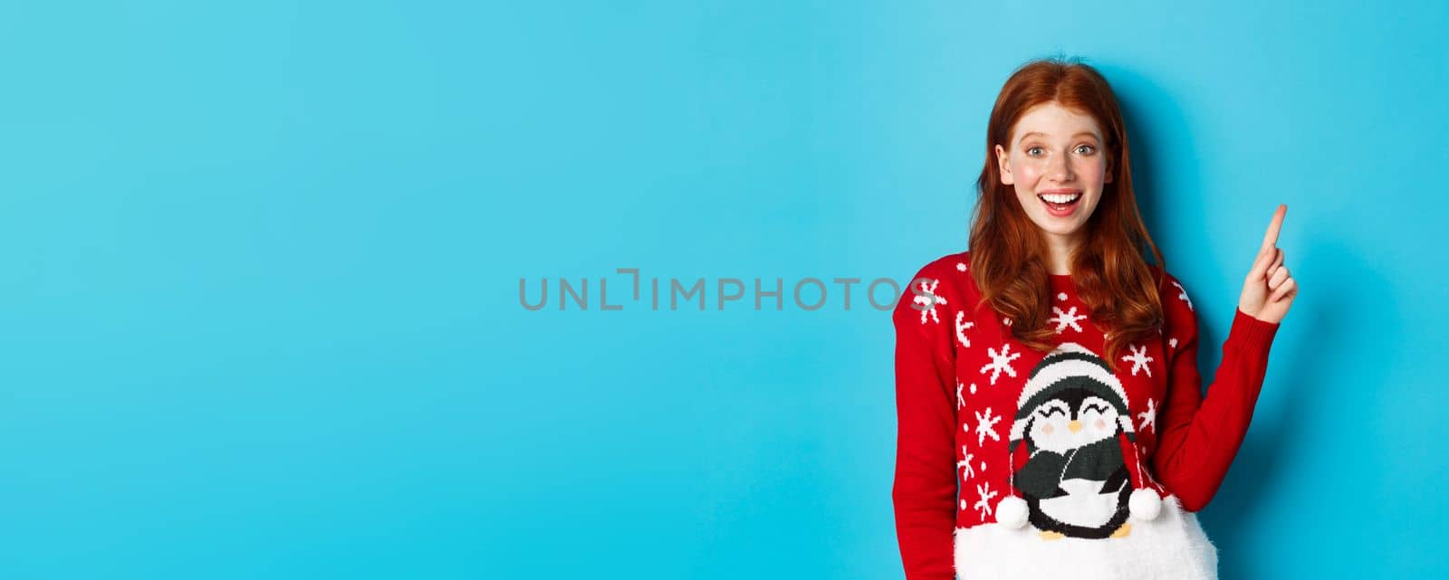 Merry Christmas. Cheerful redhead girl in xmas sweater, pointing finger at upper right corner and smiling excited, showing new year promo.