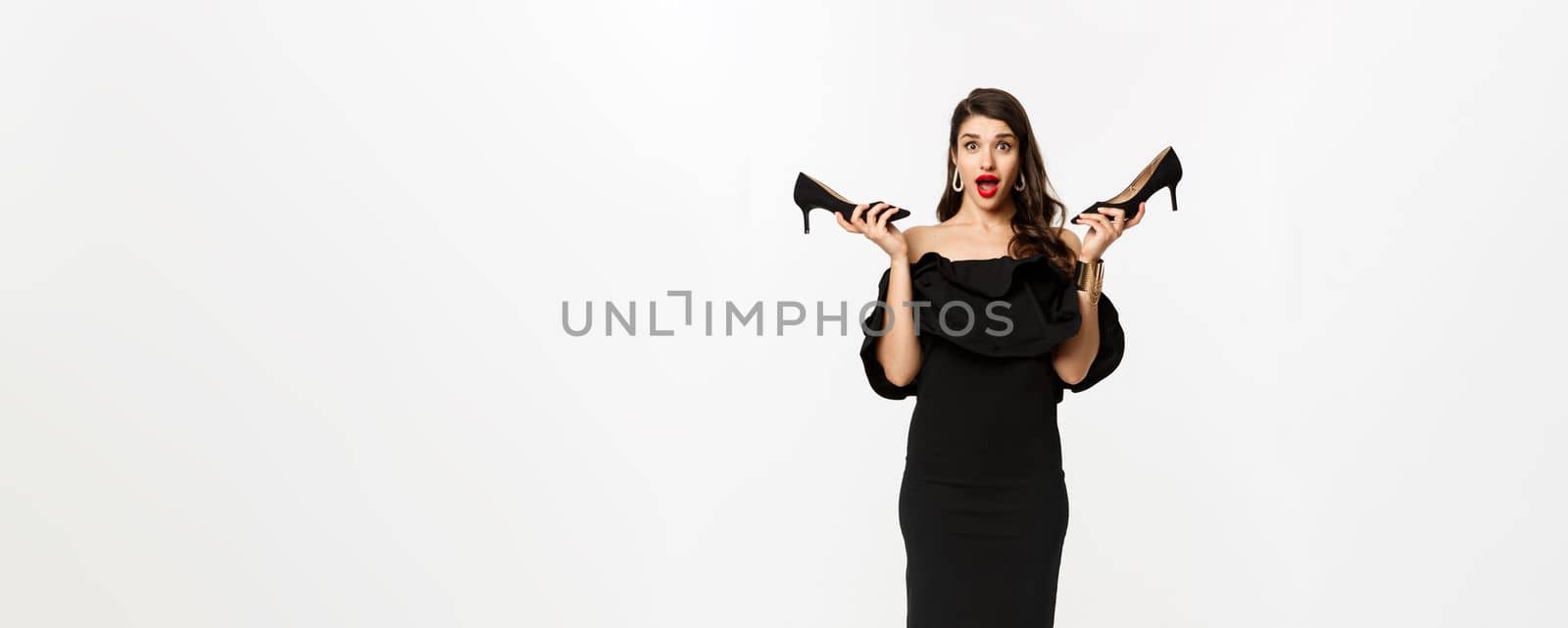 Beauty and fashion concept. Full length of excited glamour woman in black dress, showing high heels and looking excited, dressing up for party, white background.
