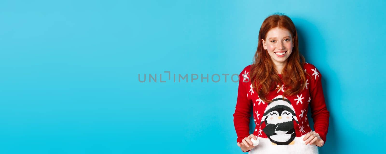 Winter holidays and Christmas Eve concept. Cute redhead girl wearing adorable sweater and smiling, celebrating New Year, standing over blue background by Benzoix
