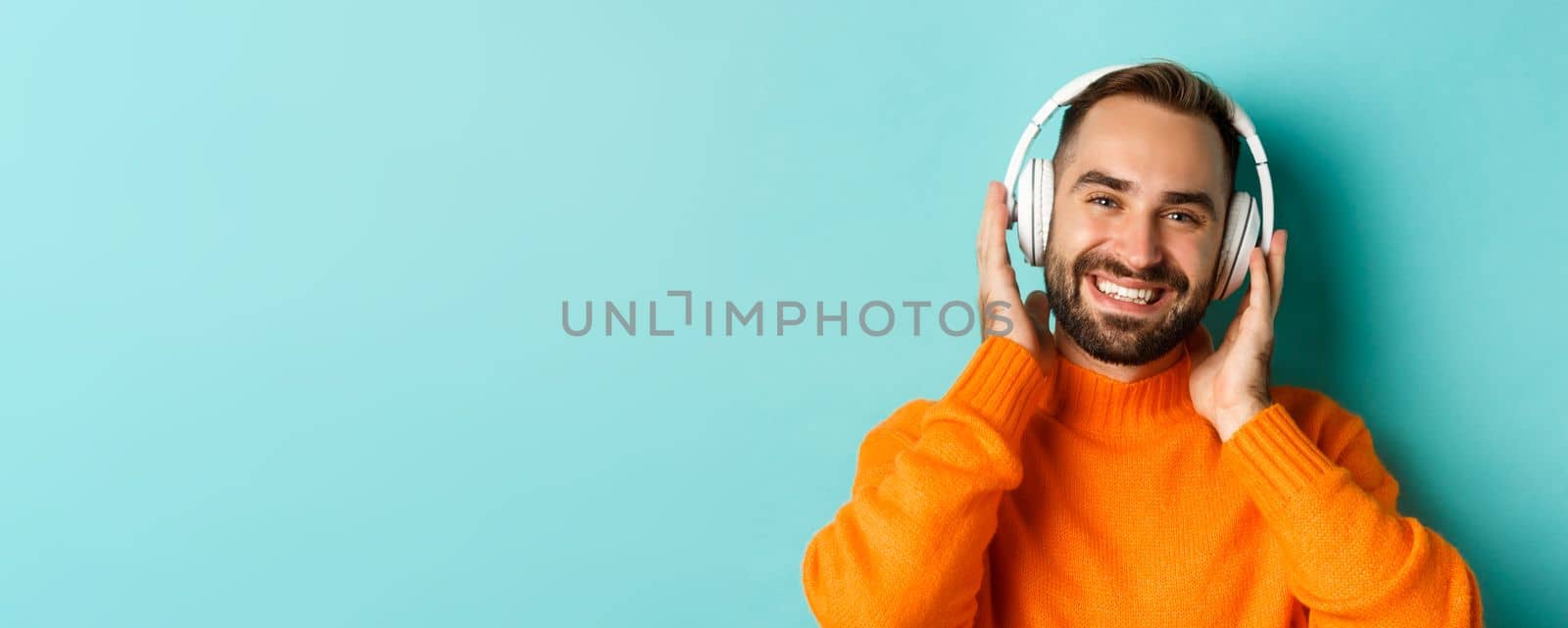 Close-up of handsome modern man listening music in headphones, standing in orange sweater over turquoise background.