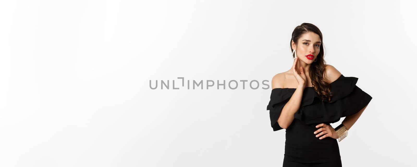 Sensual young woman in black dress, showing her earrings and looking sexy at camera, standing over white background.