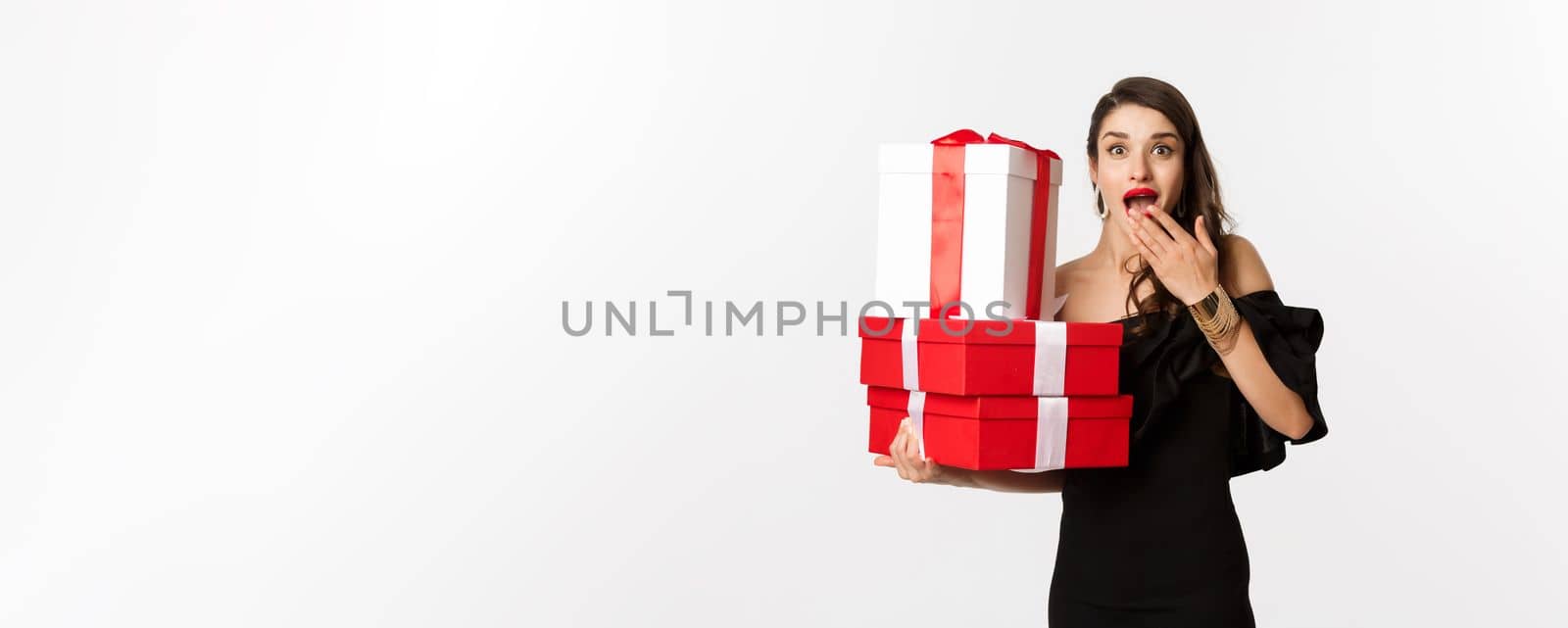 Celebration and christmas holidays concept. Woman holding xmas gifts and looking surprised, receive presents, standing over white background.