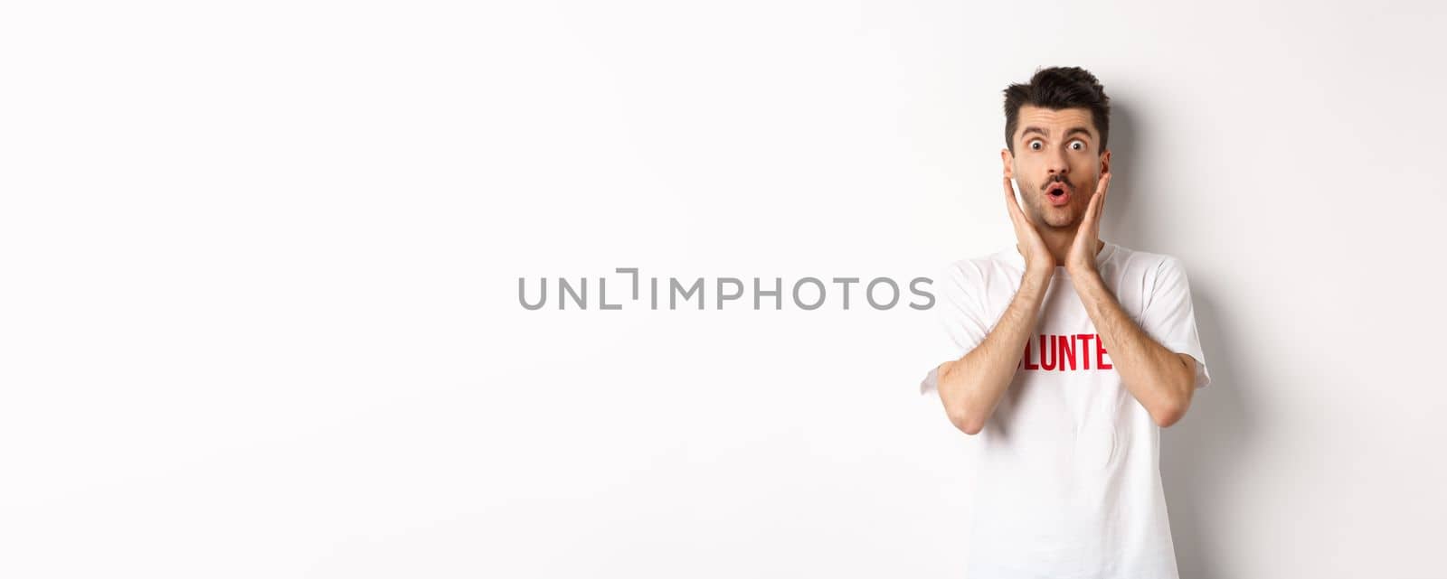 Shocked man in volunteer t-shirt gasping, staring at camera in awe, standing over white background.