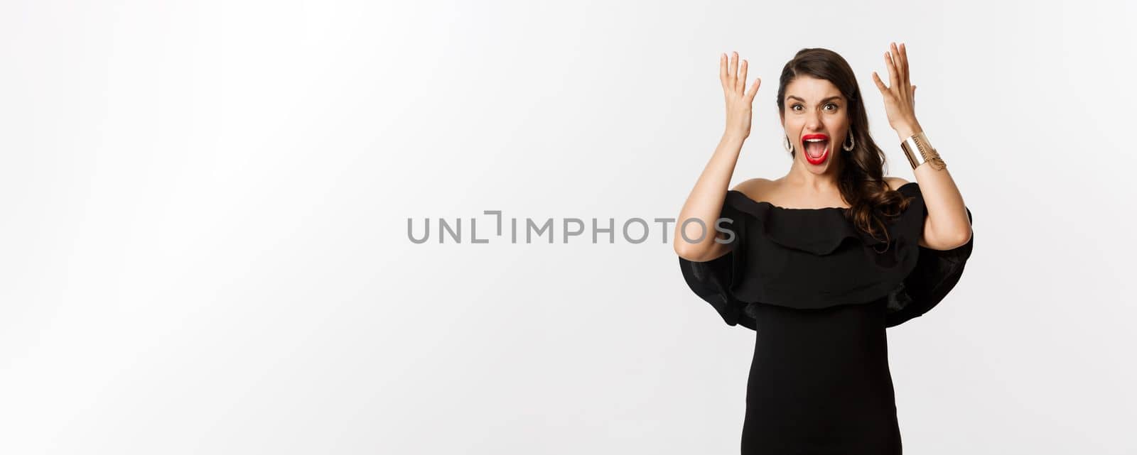 Fashion and beauty. Angry woman in black dress, shouting mad and shaking hands, grimacing outraged at camera, standing over white background by Benzoix