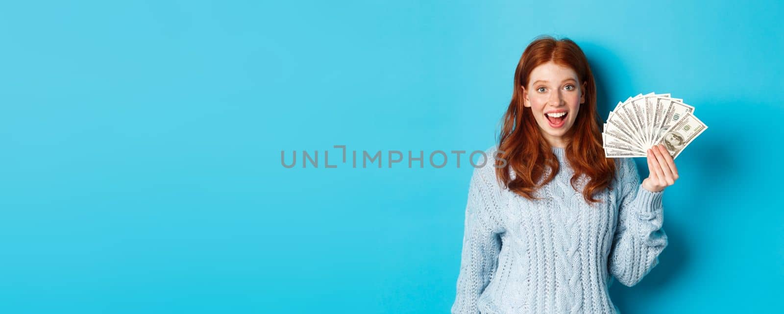 Happy redhead girl in sweater, staring excited at camera, showing money dollars, standing against blue background by Benzoix