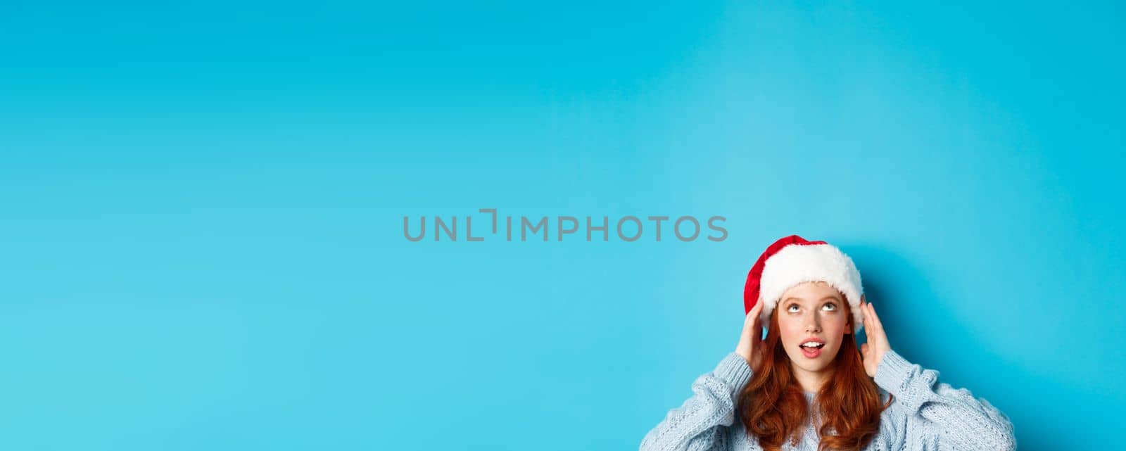 Winter holidays and Christmas eve concept. Head of cute redhead girl in santa hat, appear from bottom and looking up at copy space, staring logo, standing over blue background.