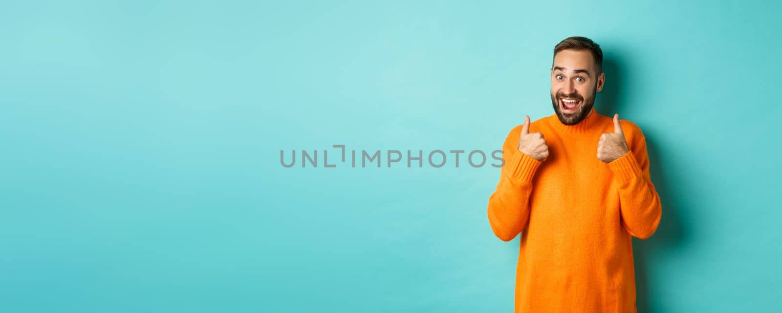 Excited young man with beard, showing thumbs up in approval, praise or recommend, standing over light blue background.