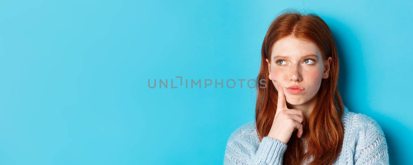 Headshot of troubled teenage girl thinking, looking bothered and frowning, standing against blue background.