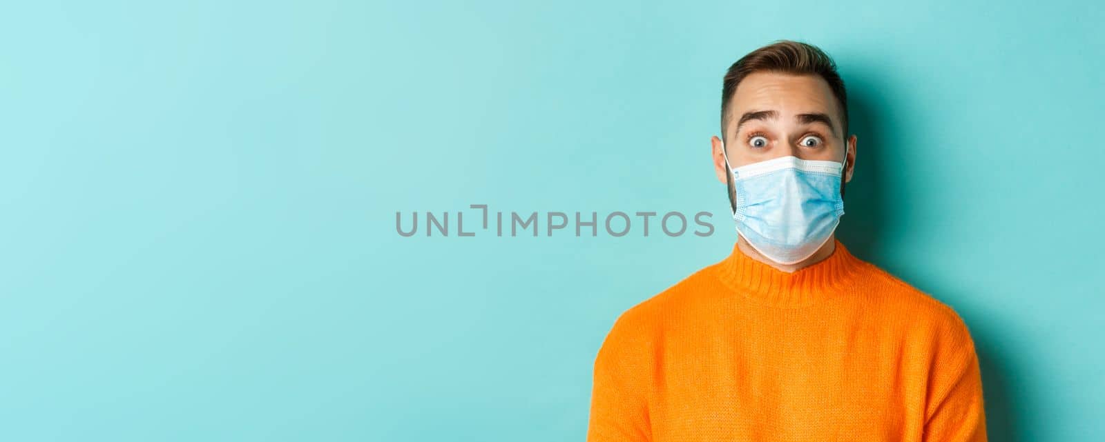 Covid-19, social distancing and quarantine concept. Close-up of young man in face mask staring at camera with surprised face, standing over turquoise background.