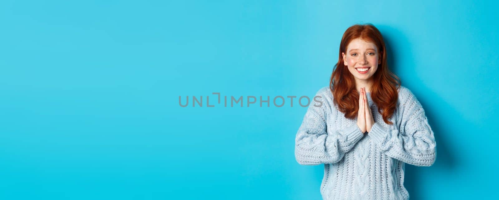 Cute redhead girl saying thank you, smiling and looking at camera, expressing gratitude, standing against blue background.