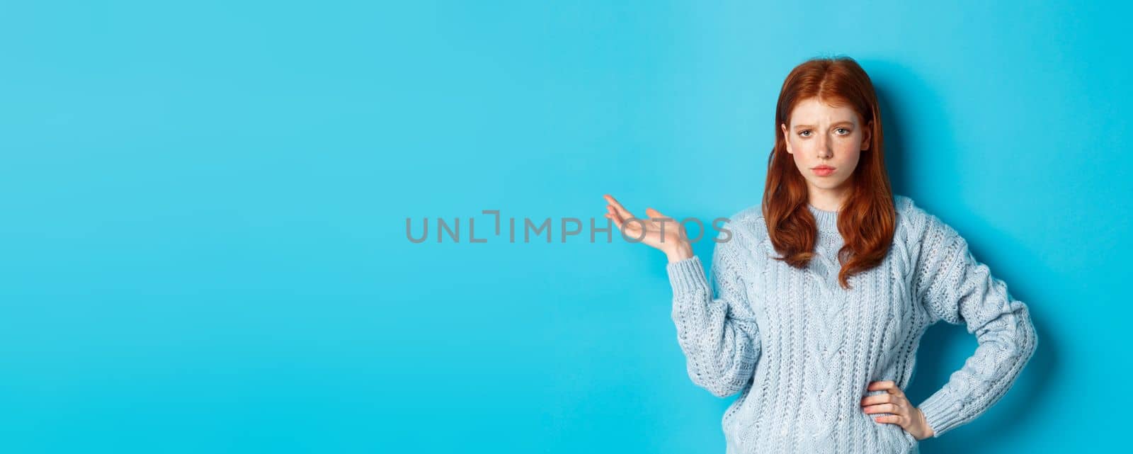 Skeptical teenage girl looking unamused, raising hand in so what gesture, staring at something with careless face, standing over blue background.