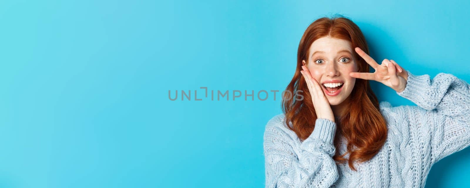 Close-up of beautiful smiling girl with red hair, showing peace kawaii sign and gazing at camera, standing over blue background.