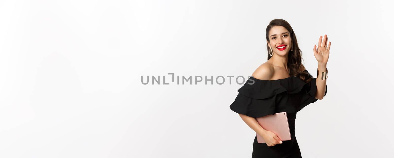 Fashion and shopping concept. Stylish young woman with glamour makeup, wearing black dress, holding digital tablet and saying hi, waving hand to greet you, white background.