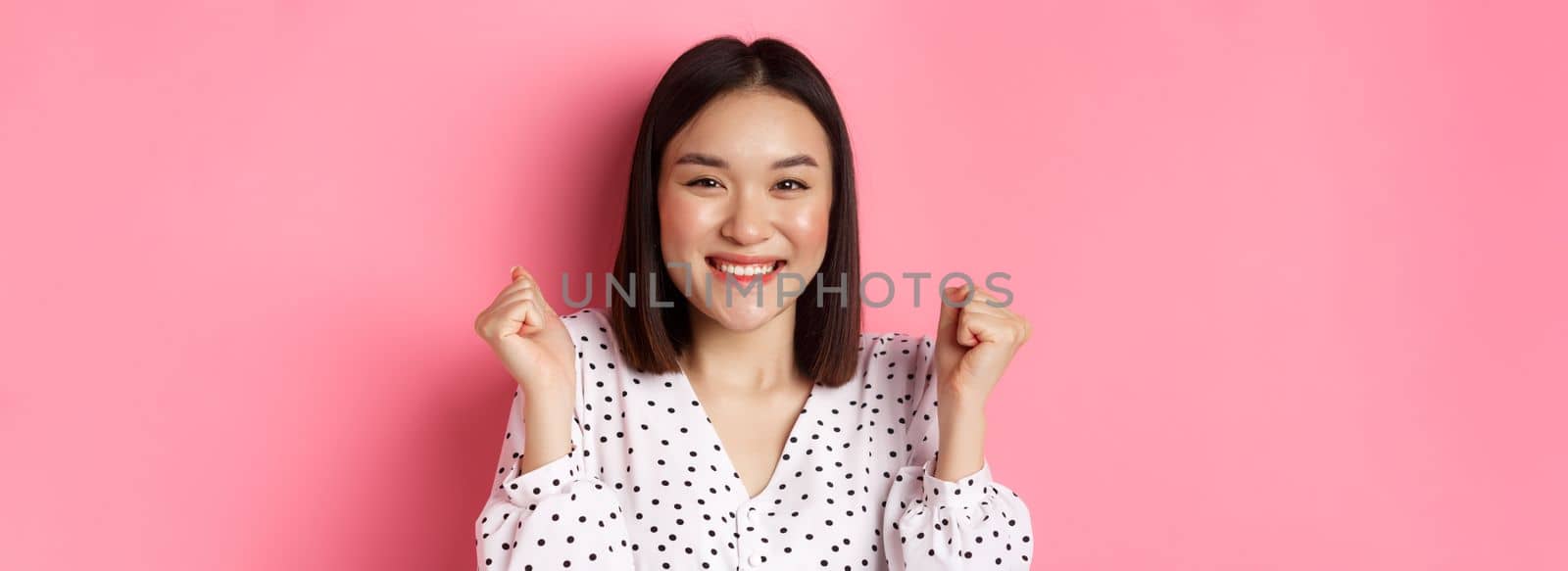 Beauty and lifestyle concept. Close-up of joyful asian woman celebrating, making fist pump and smiling of joy, winning and triumphing, standing over pink background by Benzoix