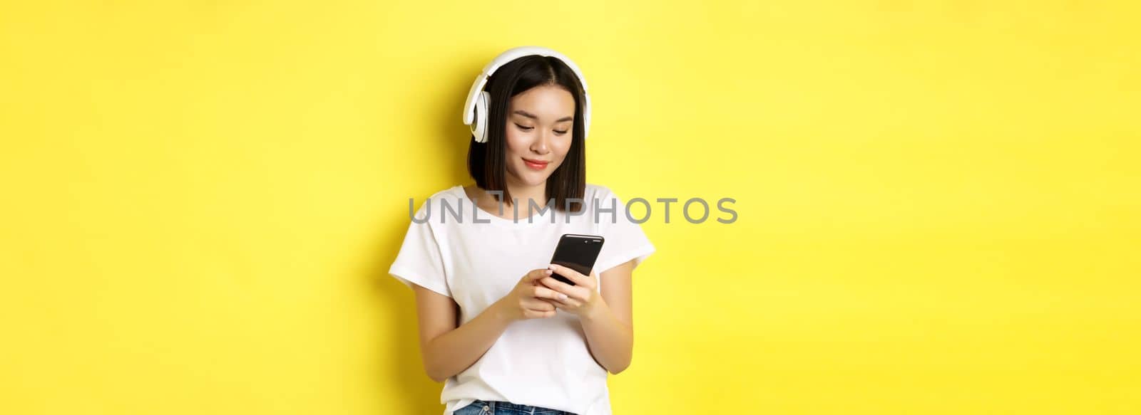 Modern asian girl listening music in wireless headphones, reading smartphone screen and smiling, standing in white t-shirt over yellow background by Benzoix