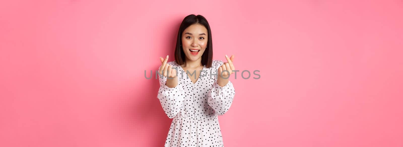 Lovely asian woman in dress showing korean heart signs and smiling, standing on romantic pink background by Benzoix