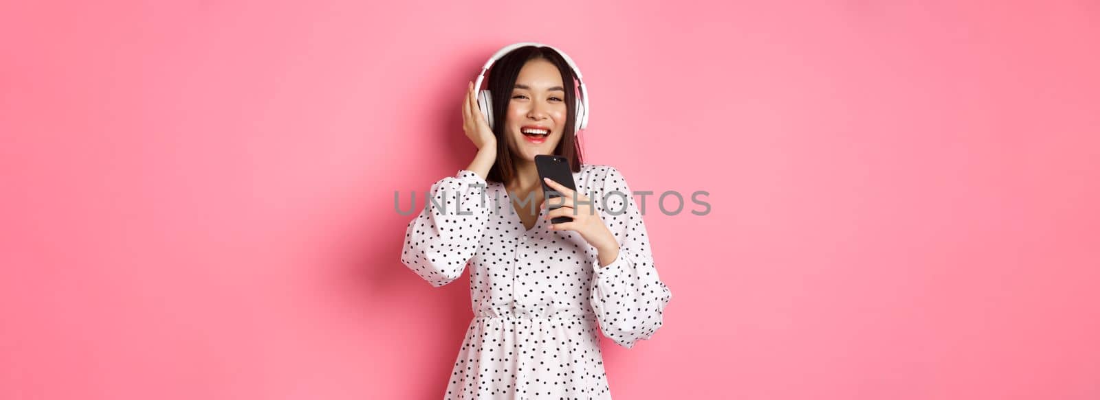 Beautiful smiling asian woman singing song in smartphone microphone, playing karaoke app and using headphones, standing over pink background.
