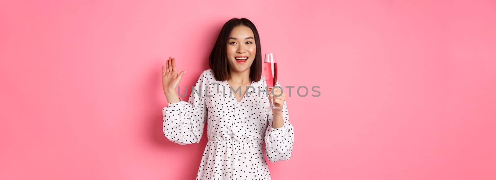 Celebration. Beautiful asian woman drinking champagne and smiling, standing in dress over pink background by Benzoix