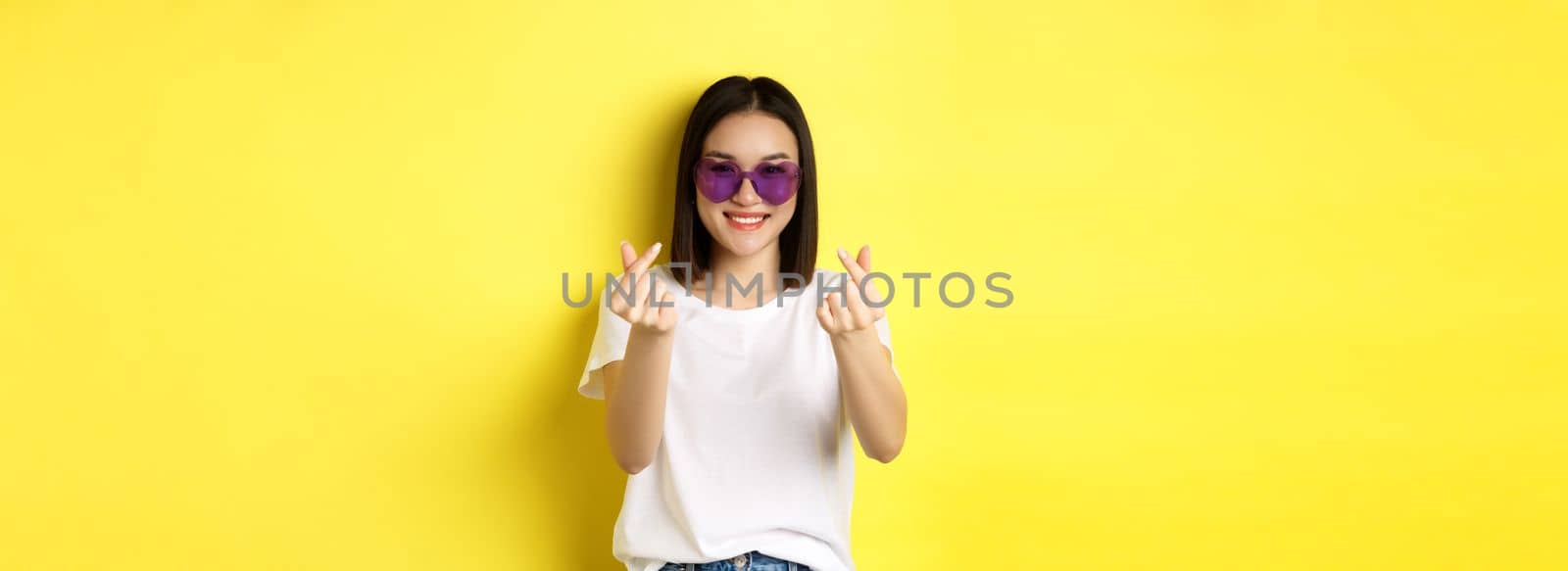 Fashion and lifestyle concept. Attractive asian woman in heart-shape sunglasses, showing finger hearts and smiling happy at camera, standing over yellow background.
