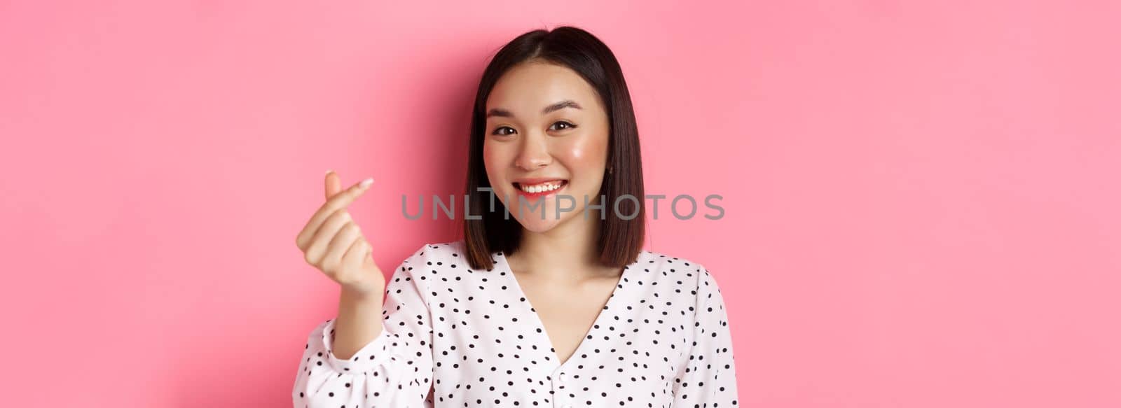 Beauty and lifestyle concept. Close-up of lovely asian woman showing heart sign, smiling and feeling romantic on valentines day, standing over pink background by Benzoix