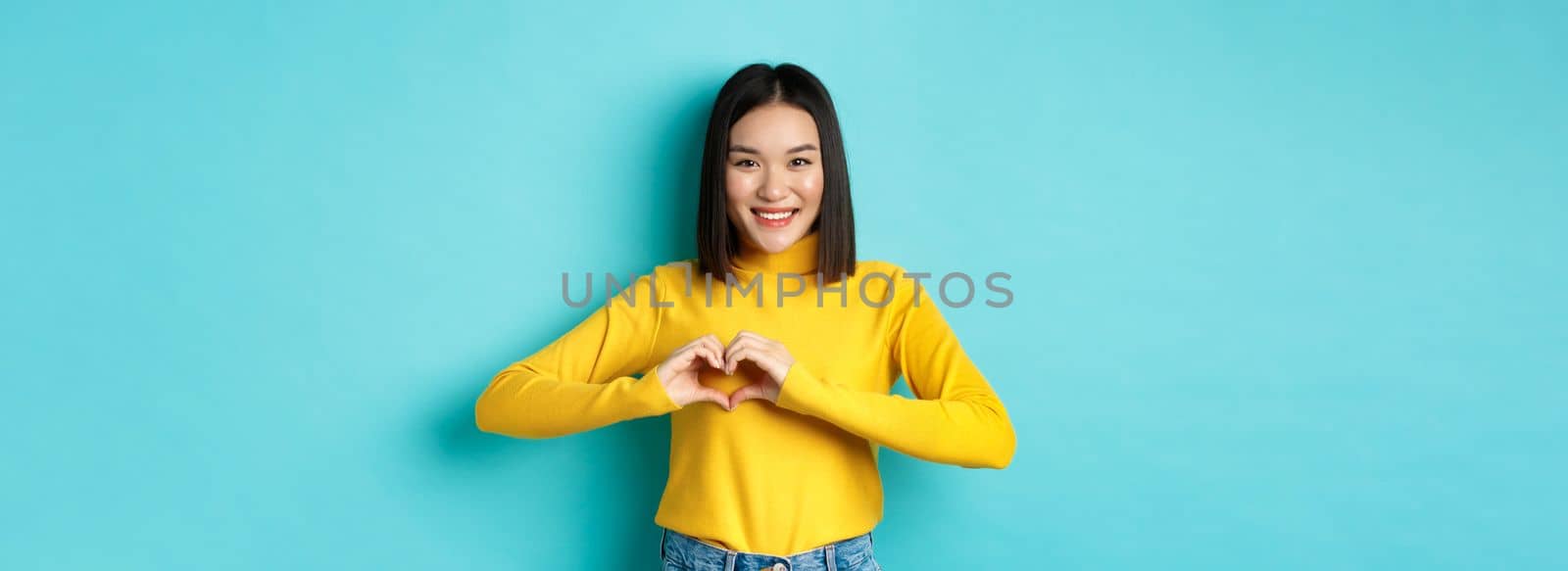 Valentines day and romance concept. Beautiful asian woman show I love you, heart gesture and smiling, standing against blue background by Benzoix