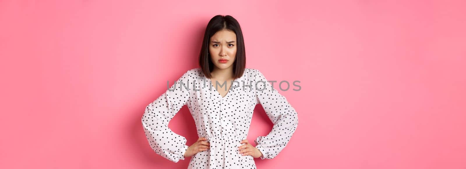 Cute offended girl frowning, holding hands on waist and staring at camera disappointed, standing upset and gloomy over pink background.