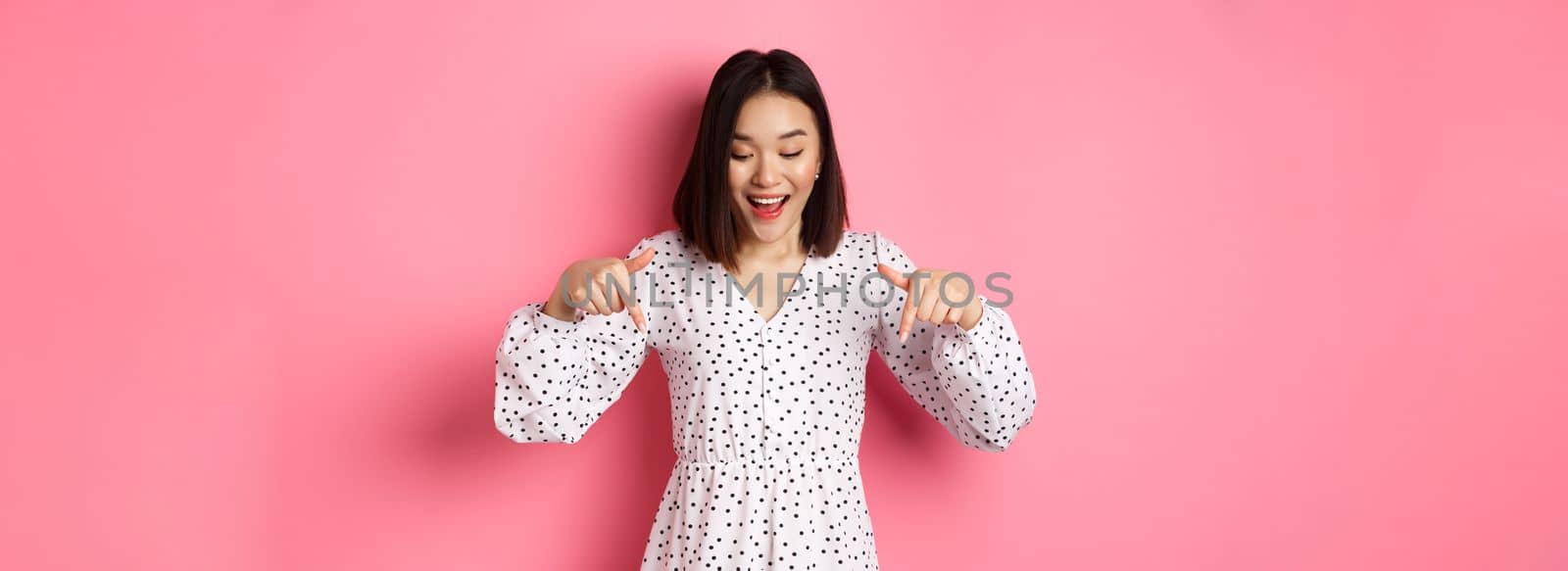 Romantic asian woman peeking down, pointing fingers at bottom, looking curious at product discount with happy smile, standing over pink background by Benzoix