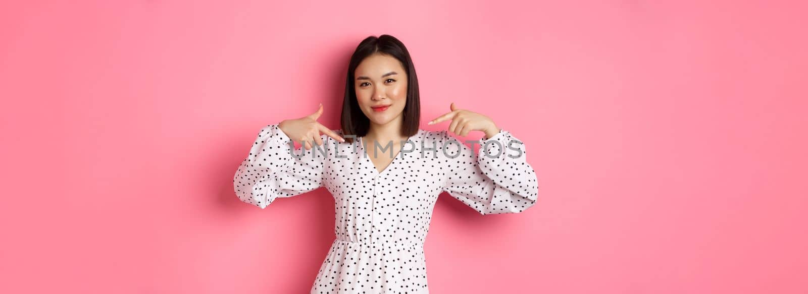 Beautiful asian woman looking confident, pointing at herself and smiling, showing logo on chest, standing over pink background by Benzoix