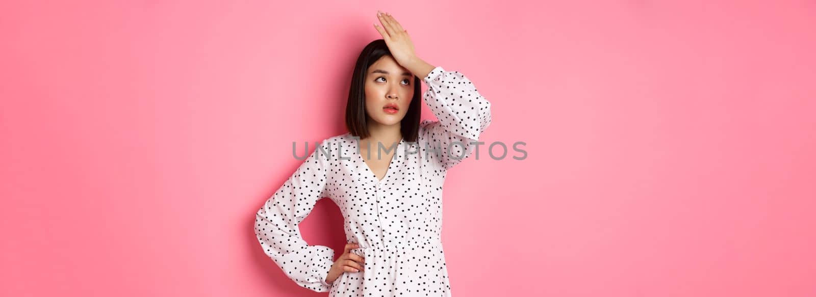 Annoyed and tired asian woman making facepalm, slap forehad and roll eyes disappointed, standing in white dress against pink background.