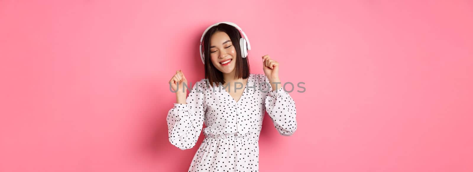 Happy young asian woman dancing and having fun, listening music in headphones, standing over pink background by Benzoix