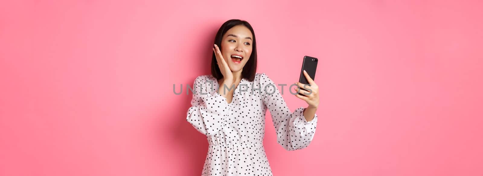 Beautiful asian girl using photo filters app and taking selfie on smartphone, posing in cute dress against pink background.