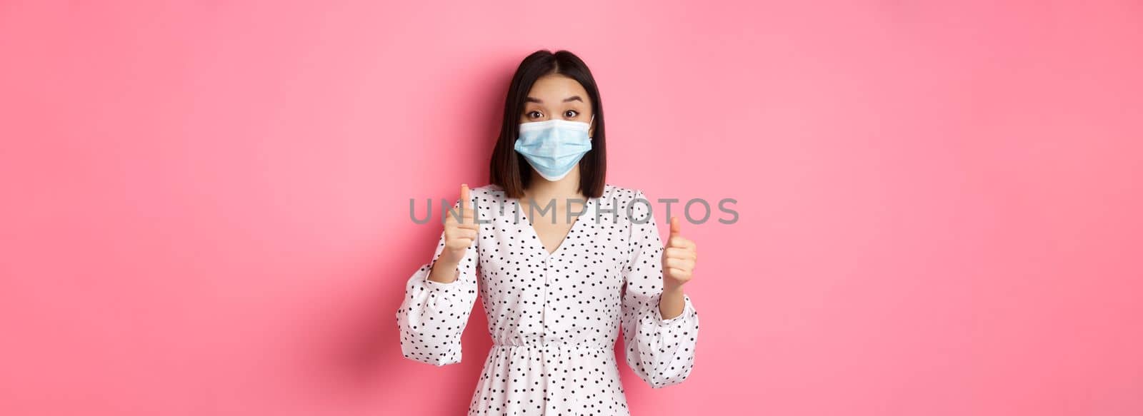 Covid-19, quarantine and lifestyle concept. Excited asian woman in face mask showing thumbs-up, praising good job, looking amazed at camera, standing over pink background.