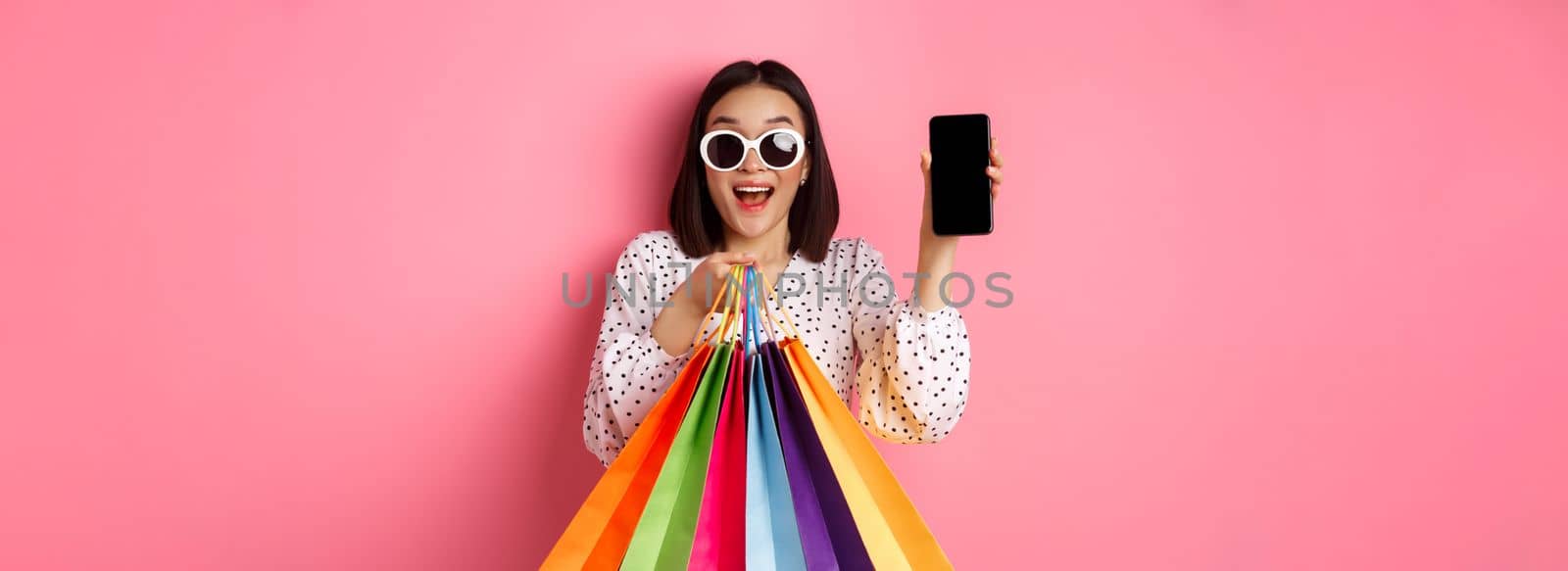 Attractive asian woman showing smartphone app and shopping bags, buying online via application, standing over pink background by Benzoix