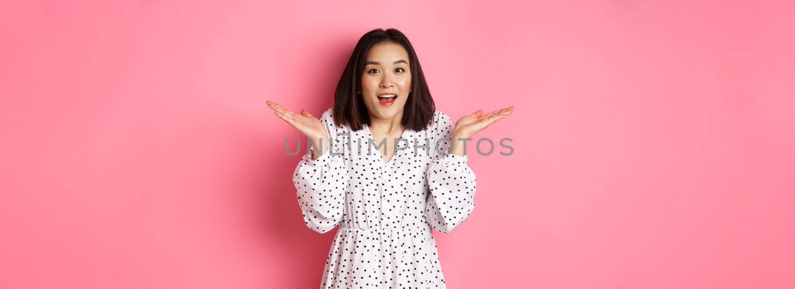 Image of surprised korean girl in dress, female model staring at camera and gasping amazd, standing over pink background.