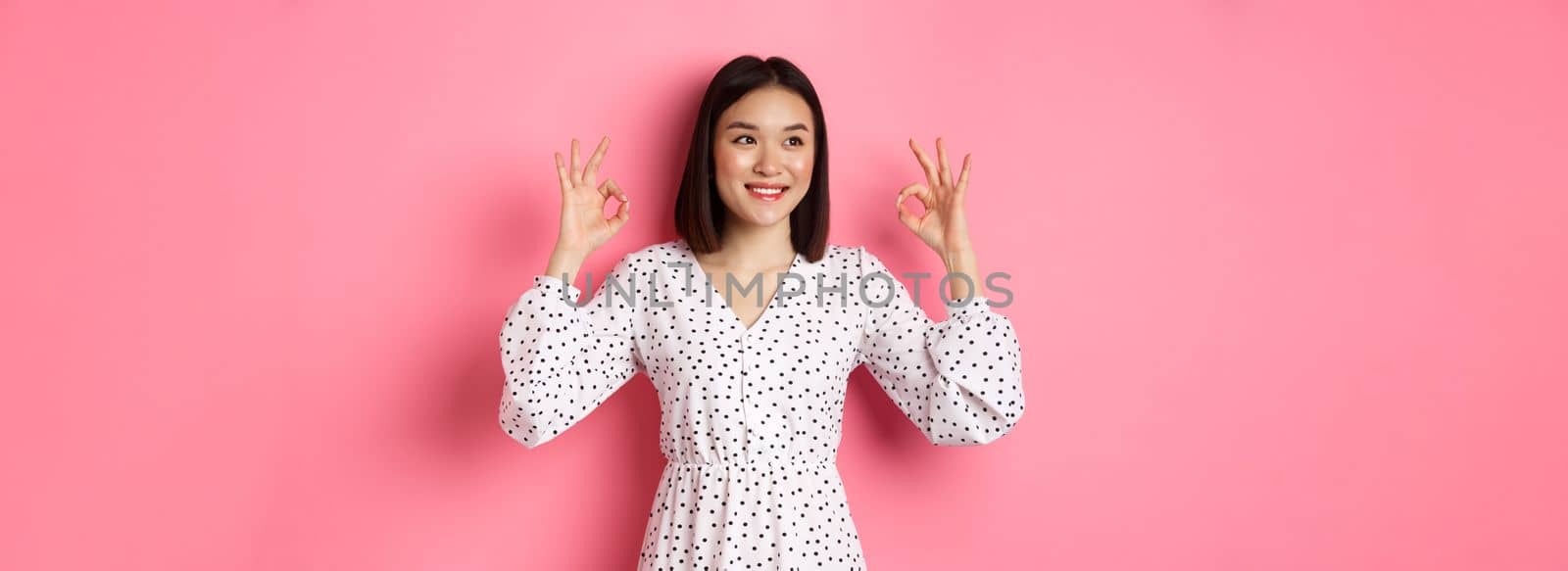 Pretty young asian woman in dress showing okay sign, praising and showing approval, looking satisfied, standing against pink background.