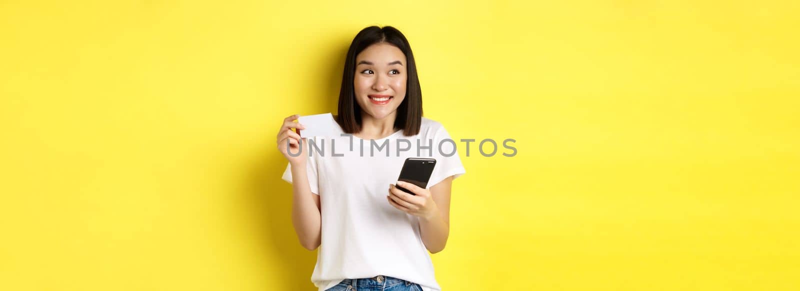 E-commerce and online shopping concept. Cheerful asian girl paying in internet, holding smartphone and plastic credit card, smiling and looking left, yellow background by Benzoix