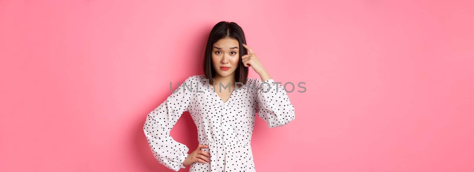 Image of attractive asian lady scolding person, pointing at head and staring at camera disappointed, are you stupid gesture, standing over pink background by Benzoix