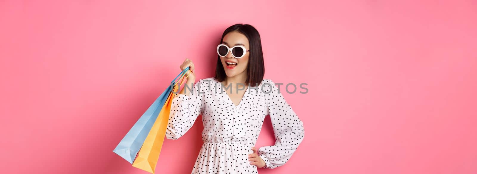 Happy modern asian woman going shopping in malls, holding bags with clothes and smiling, wearing sunglasses, standing over pink background.