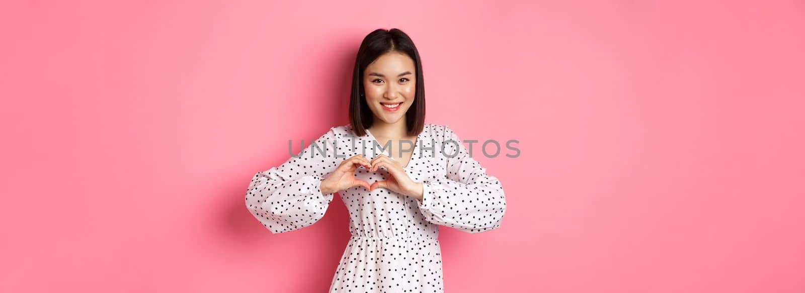 Romantic asian woman showing heart sign, I love you gesture, smiling cute at camera, standing in dress over pink background.