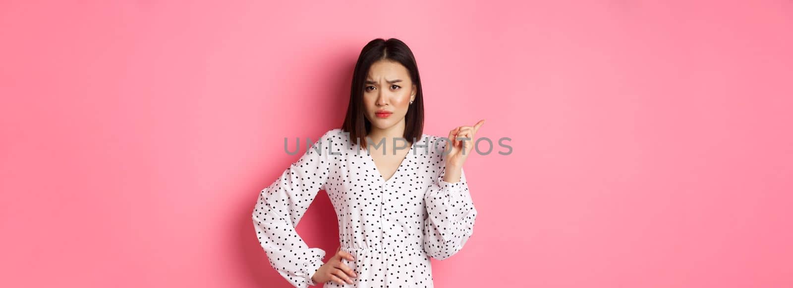 Disappointed asian girlfriend complaining, staring at camera and whining, pointing at upper left corner copy space, standing in dress over pink background.
